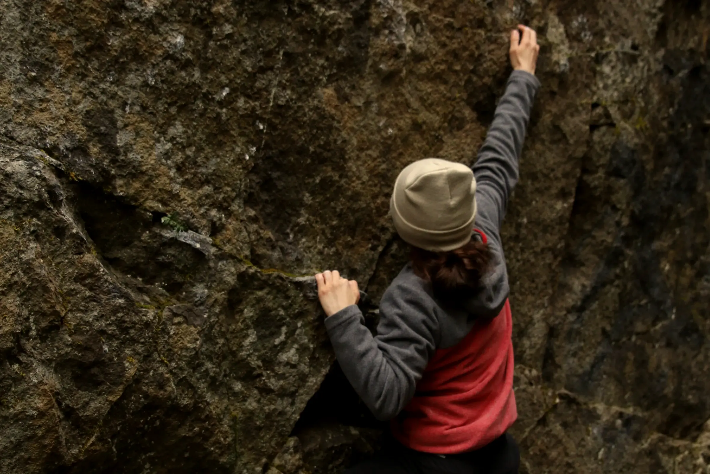 Climber in a beanie reaching up a rock face, seen from behind.