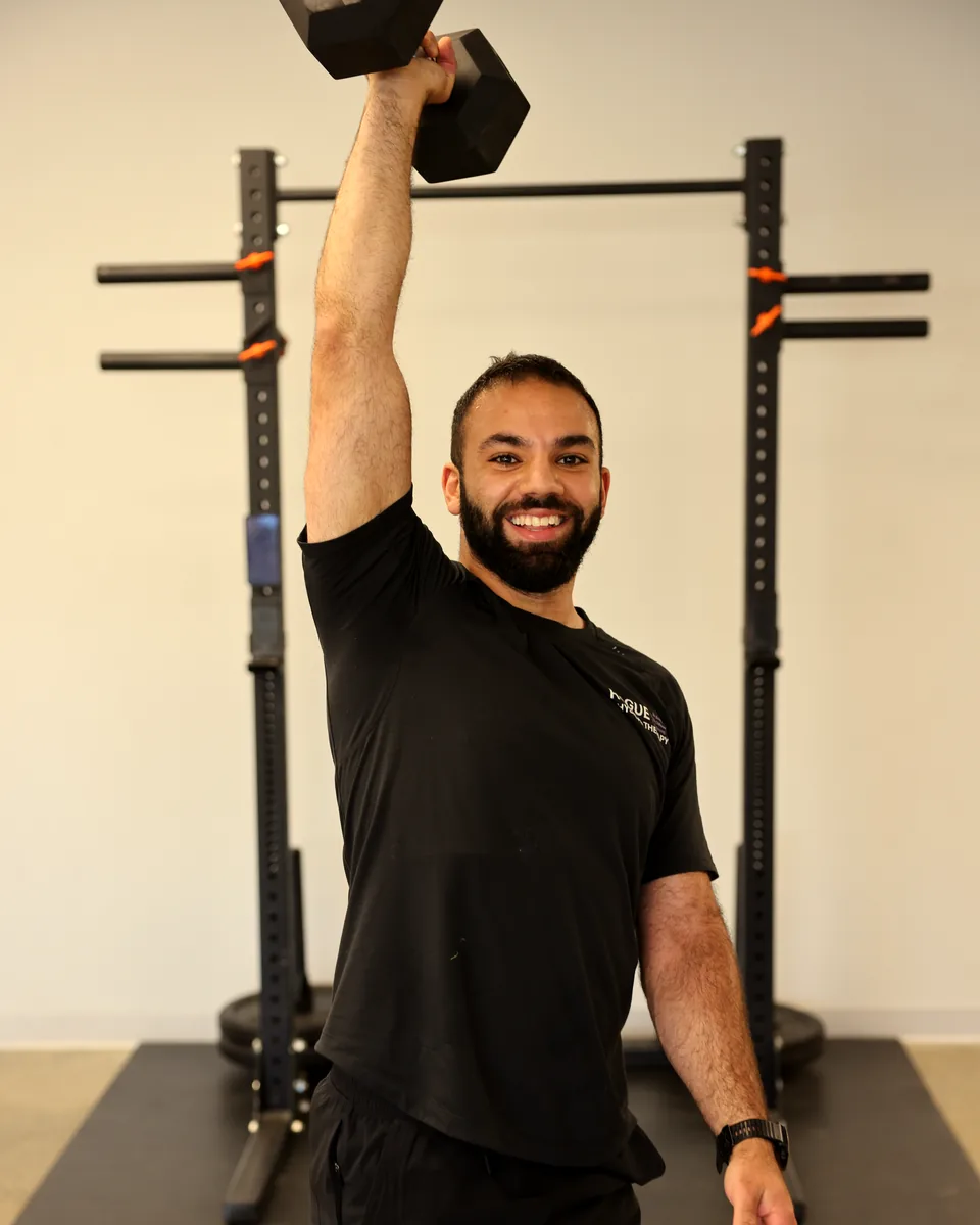 Rogue physiotherapist pressing a dumbbell overhead in front of a squat rack in the clinic gym